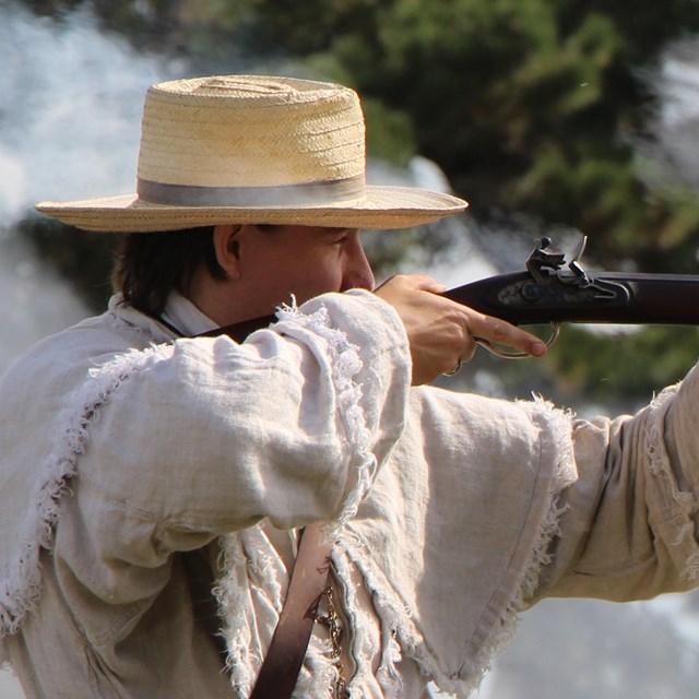 man wearing a straw hat and white overjacket shooting a rifle from era 1812