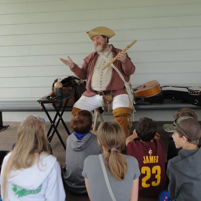 A man in a frontiersman outfit shrugs to a group of kids while holding a flute.