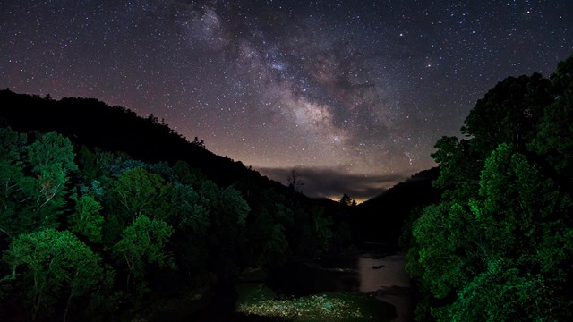 starry sky shines over orange sunset with green trees and river below