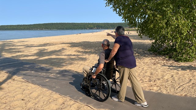 A person pushes someone in a wheelchair towards a beach on a sunny day. 