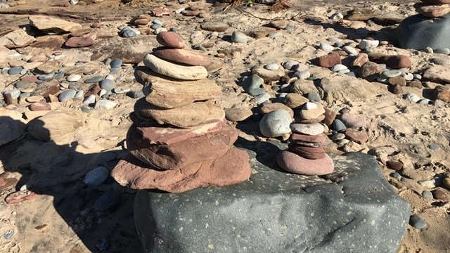Two small stacks of flat, reddish sandstone (rock cairns) sit atop a larger grey rock on a beach.