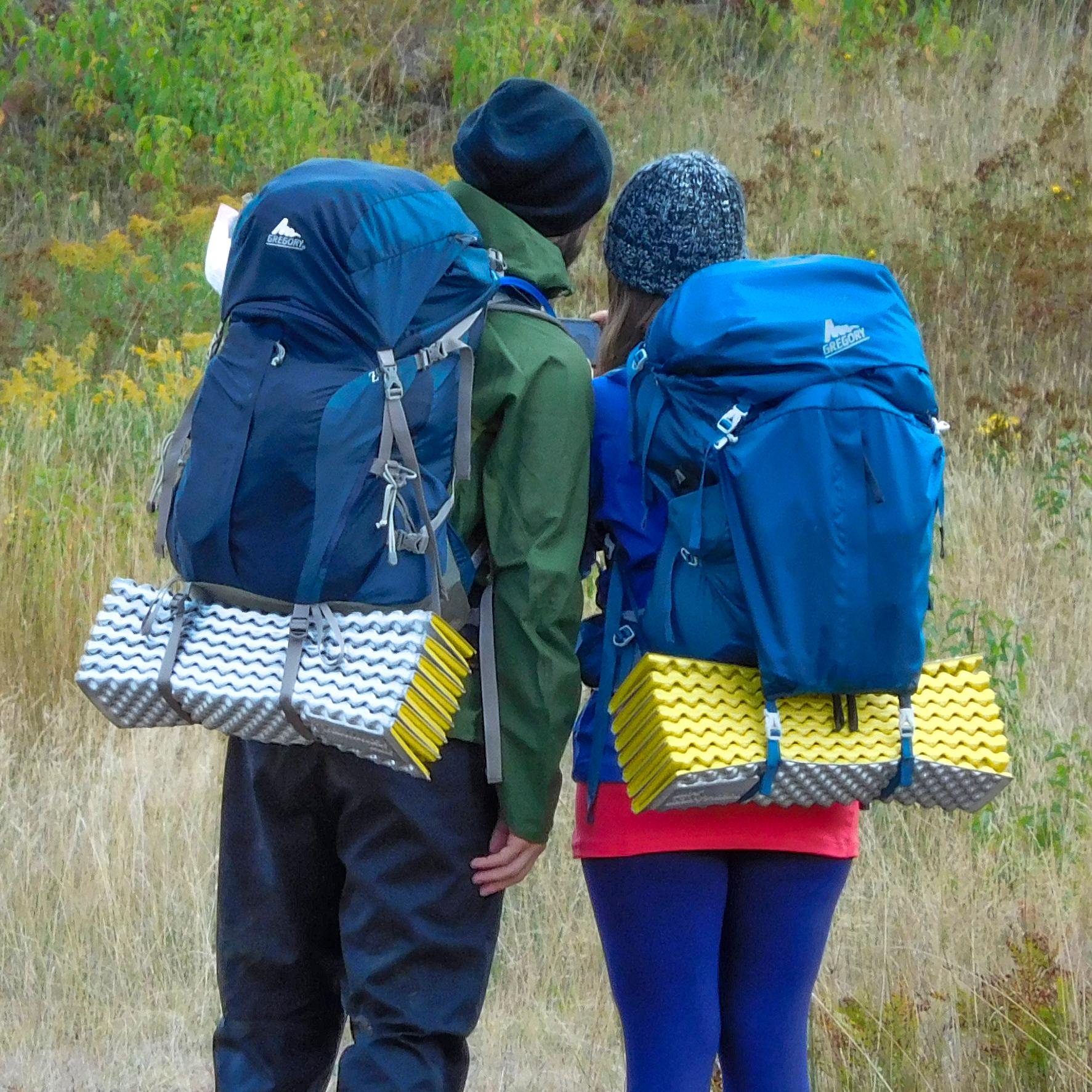 Two people with large backpacks and sleeping pads face away from the camera while looking at a map