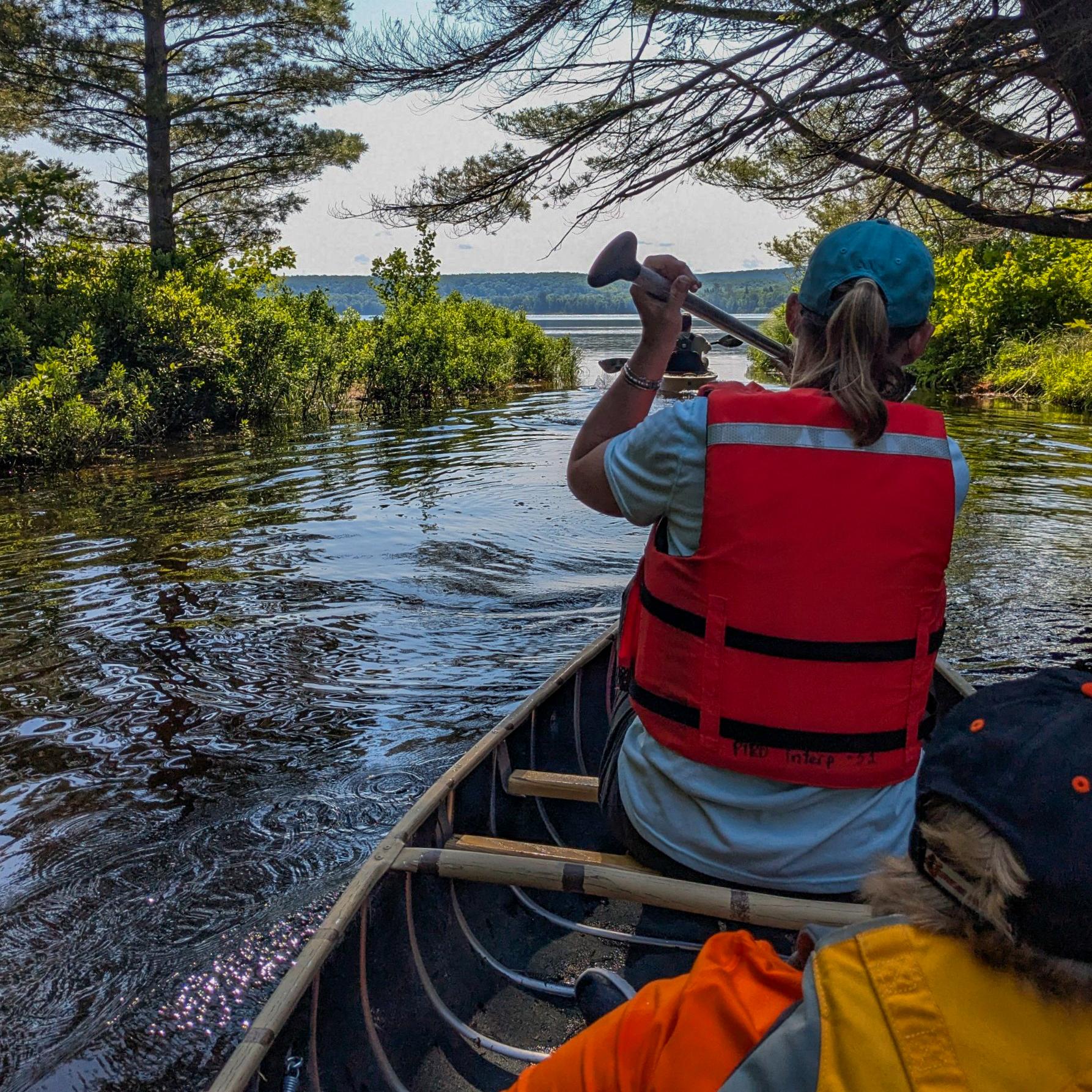 A person in a life jacket sits in the front of a canoe traveling through a channel