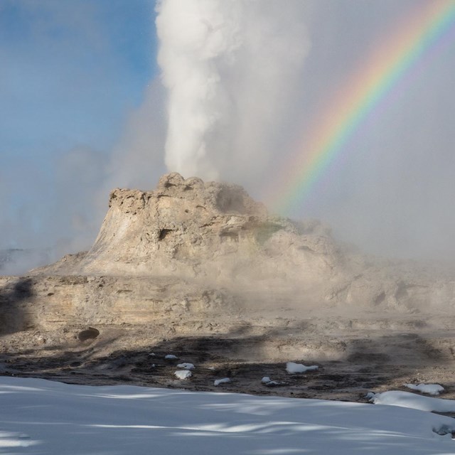 Rainbow over an erupting geyser surrounded by snow on the ground