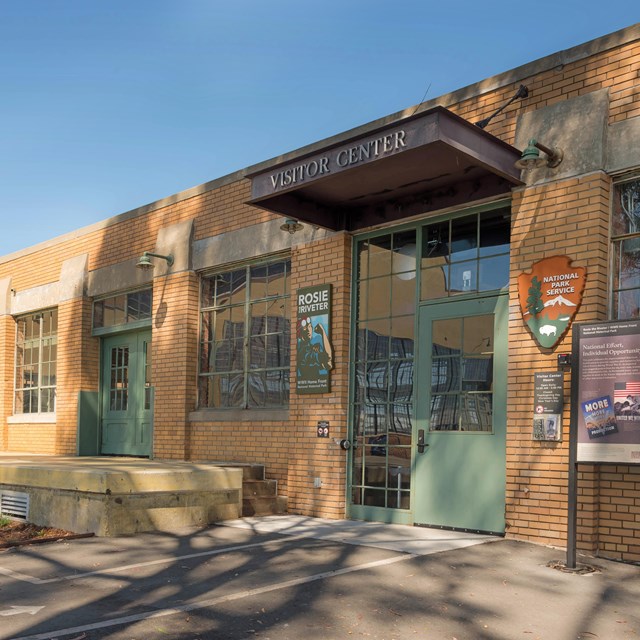 A single story brick building with many windows and a large entrance door. Signs in front. 