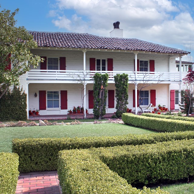 A two-story home with an upper balcony and shutters. A courtyard full of trees, shrubs and plants. 