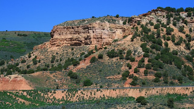 A towering red sandstone bluff, lightly vegetated, under a blue sky.