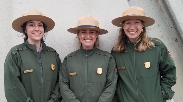 Three uniformed park rangers stand shoulder to shoulder.