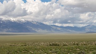 A vast grassy range leads to towering, snow-capped distant mountains, under a cloud filled sky.
