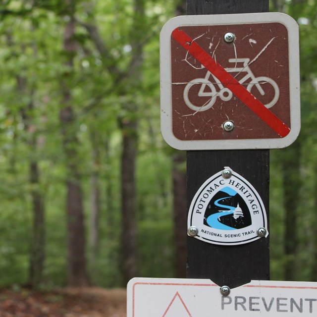A small trail logo and a no bicycle sign are attached to a wood post. In the background is a trail.