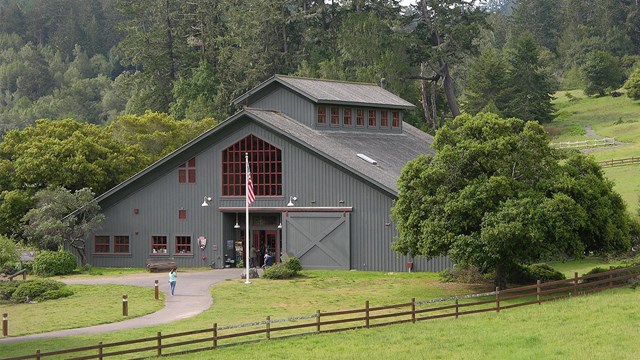 A few visitors at a gray barn-like visitor center surrounded by fenced-pasture and trees.
