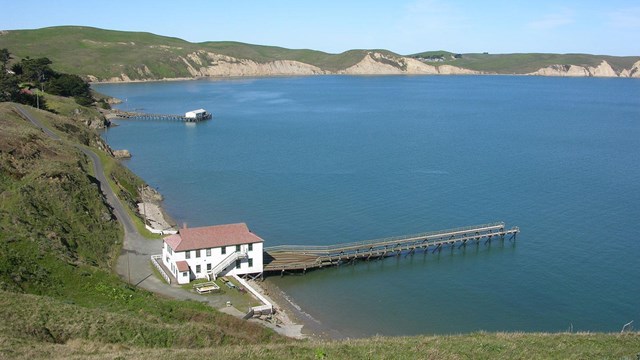 A two-story, white-sided, red-roofed building with a long dock at the edge of a blue bay.