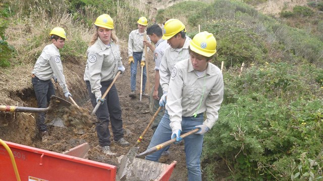 Seven youth wearing yellow hard hats and khaki-colored shirts shovel dirt from a trail.