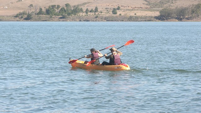 Two kayakers wearing purple PFDs paddling a yellow kayak on a calm bay.