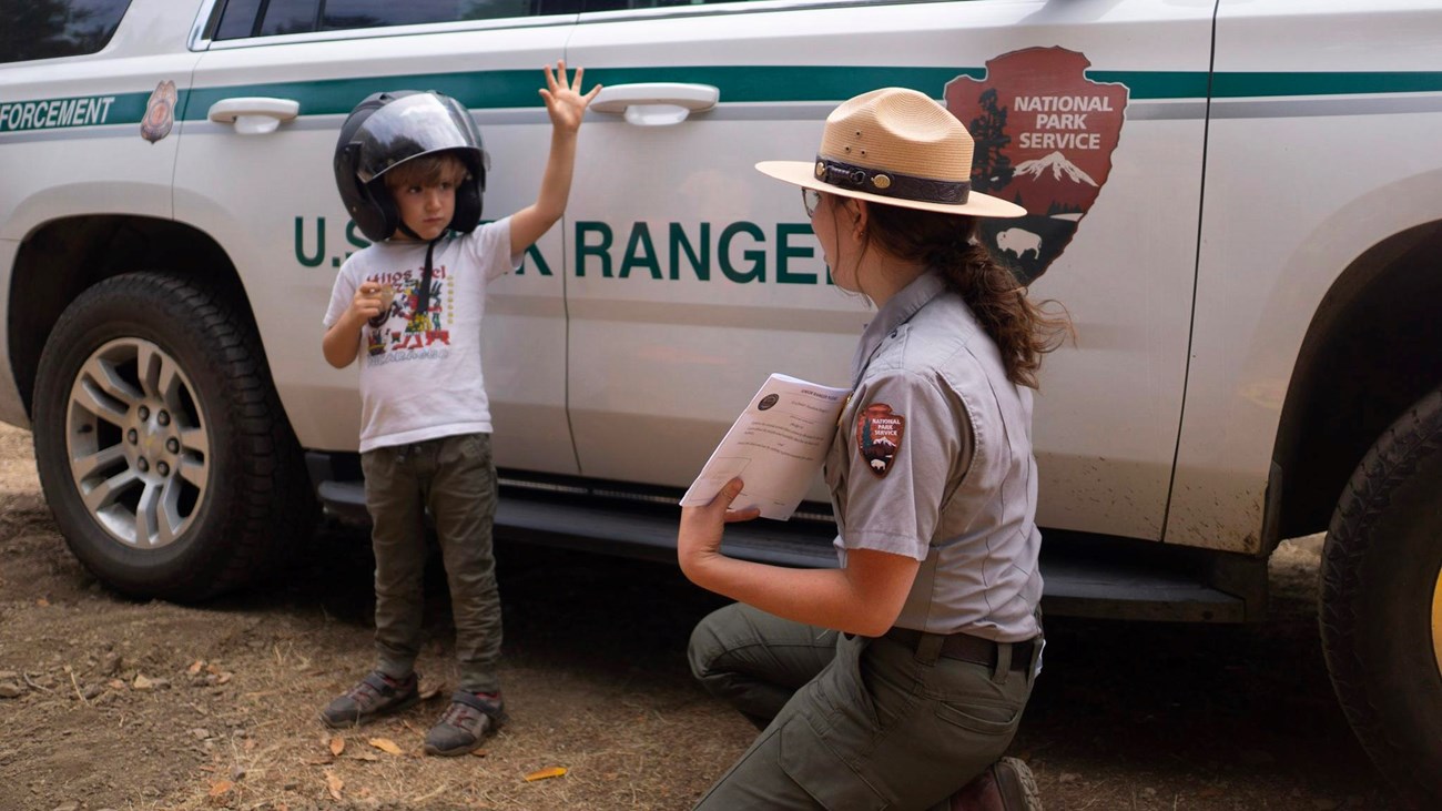 A child wearing a helmet raises his hand near a park ranger in front of a marked patrol vehicle.
