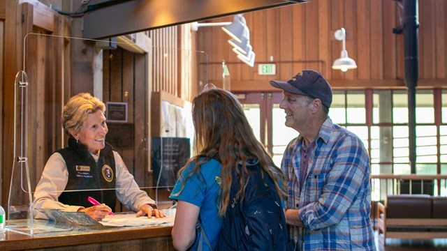 A park volunteer behind a desk talks to a man and a woman at a visitor center. 