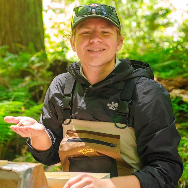 Smiling young person in a cap and waders, holding a small fish in one hand.