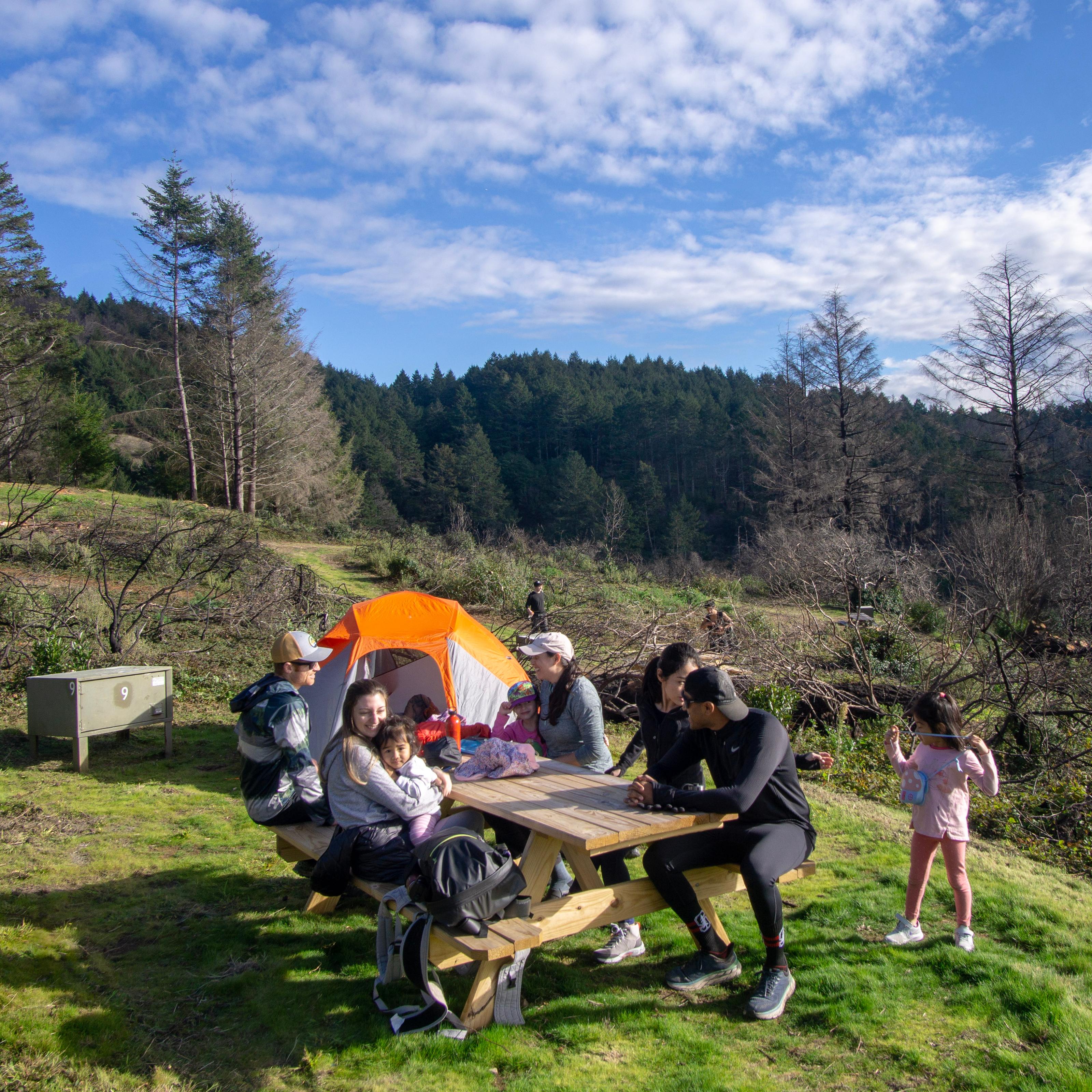 A family of seven sits around a picnic table in a campsite with a tent and food storage locker.