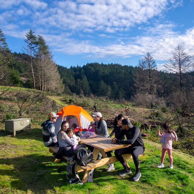 A family of seven sits around a picnic table in a campsite with a tent and food storage locker.