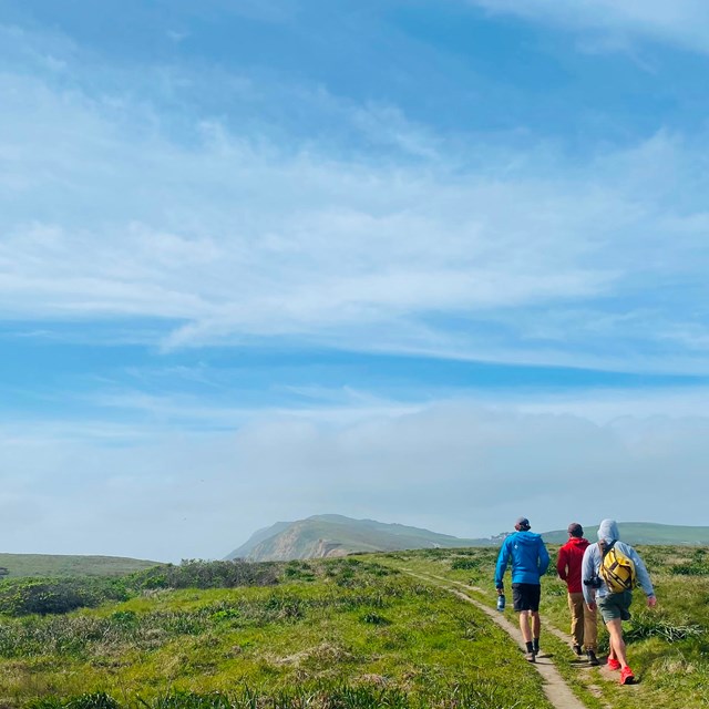 Three people hike along a dirt path through coastal grasslands under a blue partly cloudy sky.