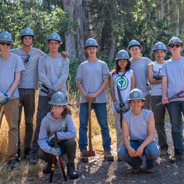 Ten teens in gray t-shirts and teal helmets stand in a forest holding hand tools. 