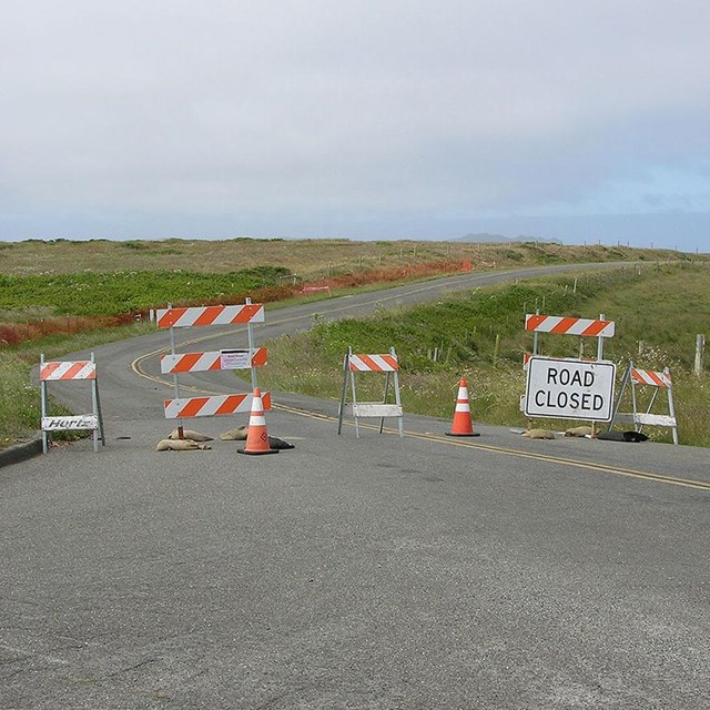 Two orange and white barricades with a road closed sign block a road through grasslands.