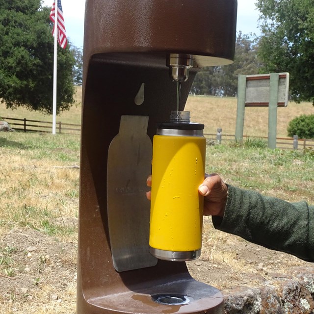 A visitor fills a yellow water bottle at a brown water bottle filling station.