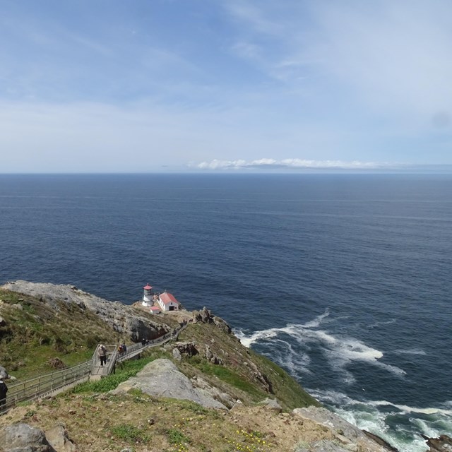 The Pacific Ocean extends beyond a small lighthouse on a rocky headland to a cloud-lined horizon.