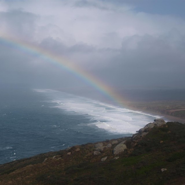 A rainbow arcs up and to the left from a beach on the right over the ocean through some clouds.