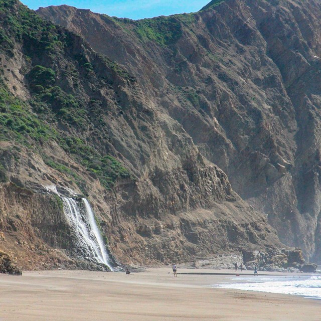 White water cascading over grass-topped coastal bluffs to a sandy ocean beach with four people below