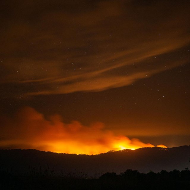 Smoke rises above a ridge and reflects orange light from a wildfire burning on the ridge's far side.
