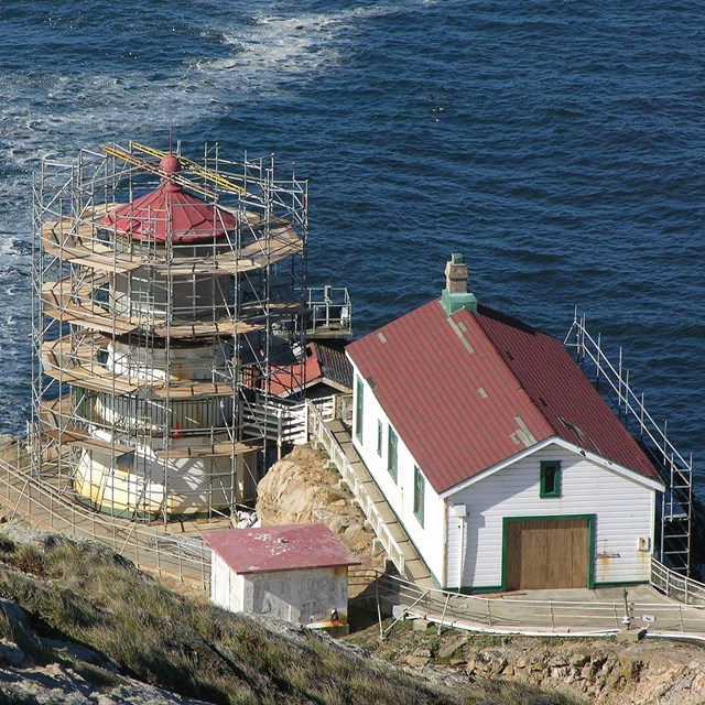 Three white-sided, red-roofed structures surrounded by scaffolding sit on an oceanic rocky headland.