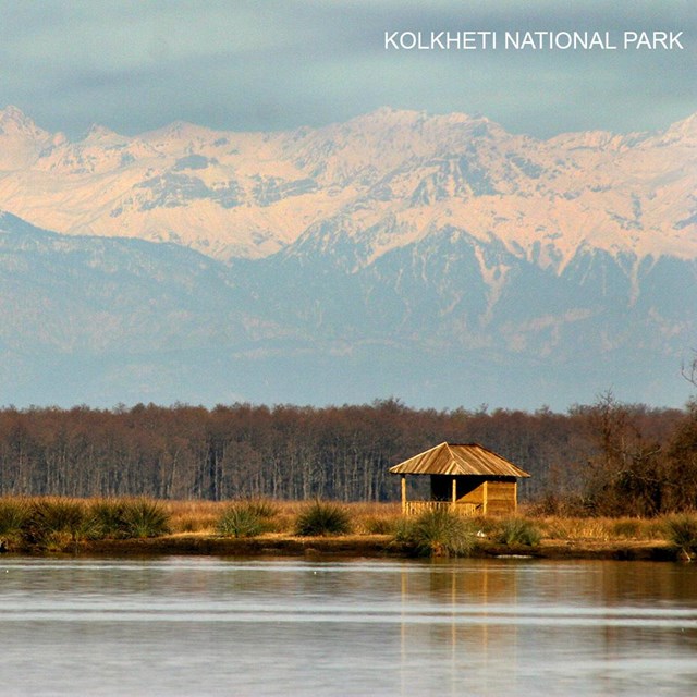 A small wooden ranger shelter on a lakeshore with a snow-capped mountain range in the background.