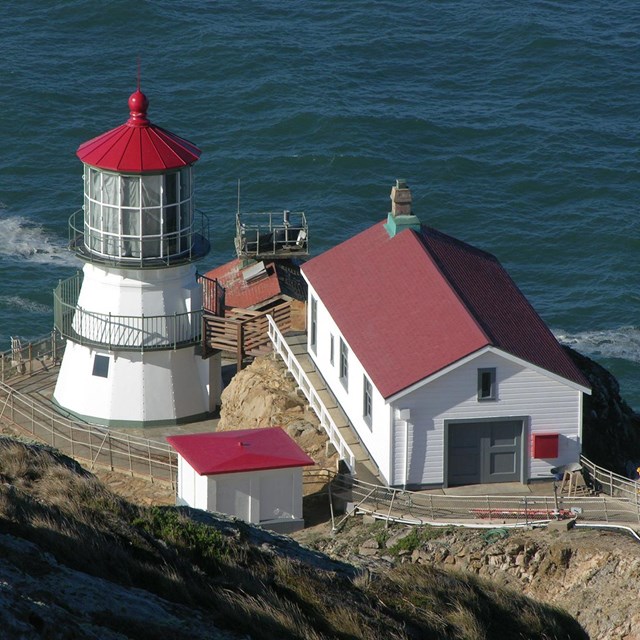 Four white-sided, red-roofed structures sit on a rocky headland above the Pacific Ocean.