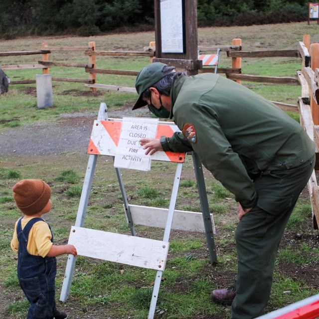 A park ranger points to a sign saying this area is closed due to fire danger, a small boy looks on.