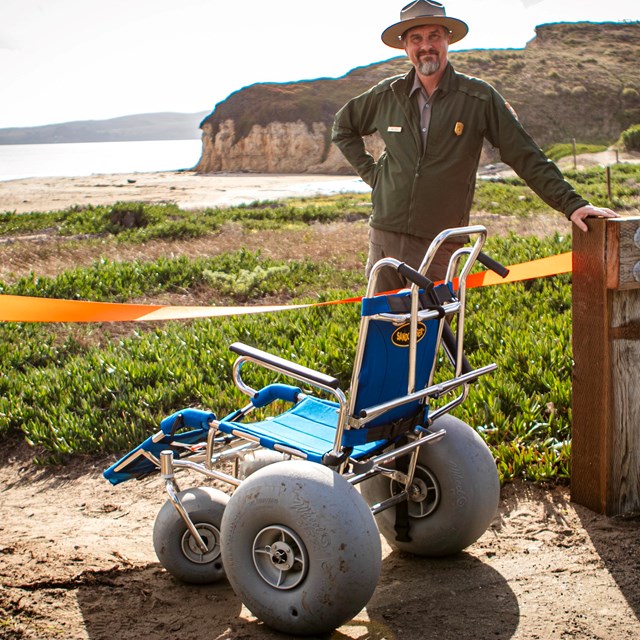 Park ranger stands smiling behind a blue wheelchair with huge gray wheels.