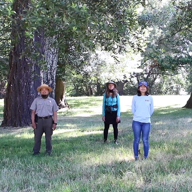 Three park rangers (one on a horse) and two women stand physically-distanced among a few trees.