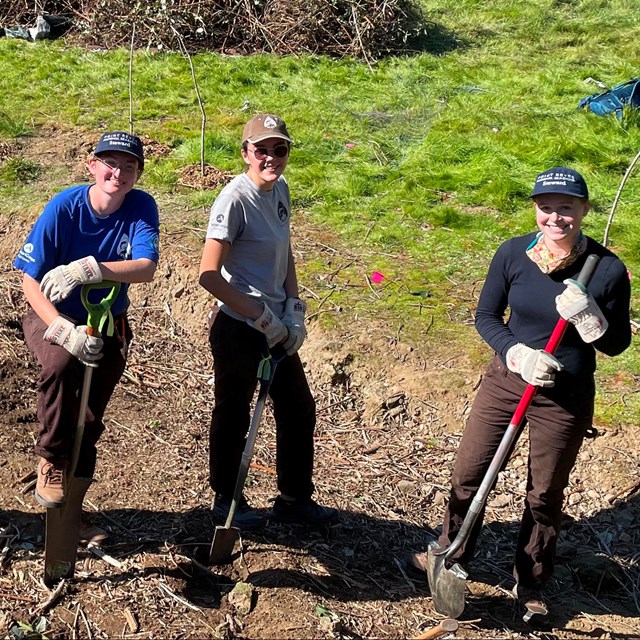 Group of people with hand tools pose outside in front of piles of plant material.