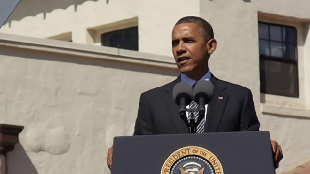 President Barak Obama stands at a podium in front of a stucco structure
