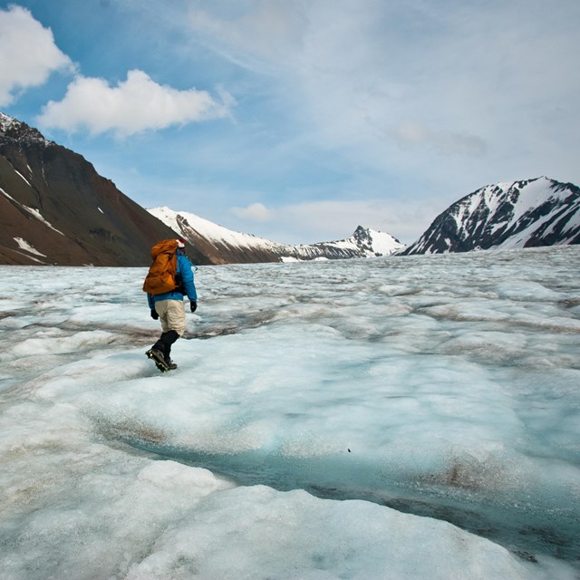 Man traverses ice field surrounded by snowy mountain peaks. 