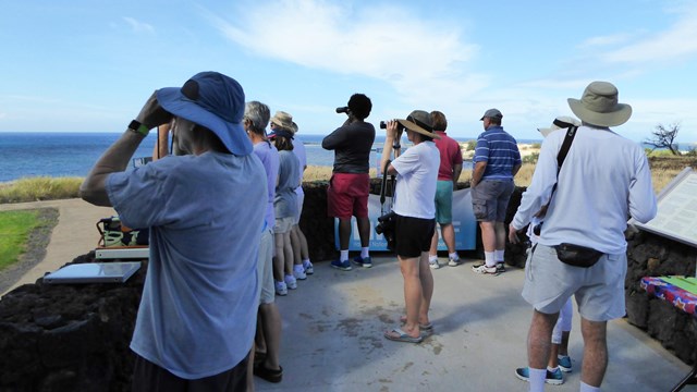Group of visitors look through pairs of binoculars toward the ocean