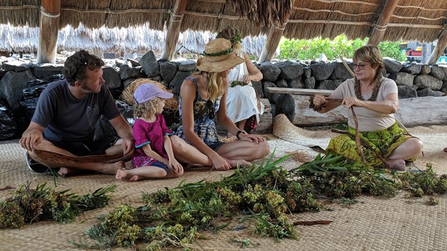 A family sitting on a lauhala mat learns how to make a lei
