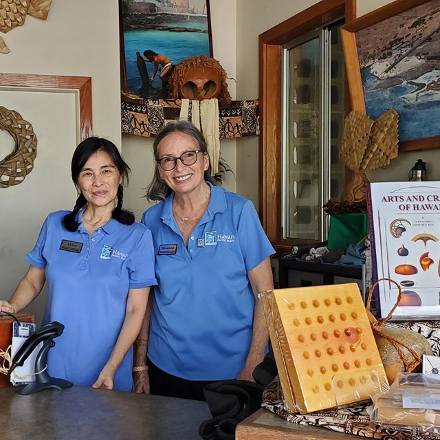 Two women in Hawaiʻi Pacific Parks Association polo shirts smile from behind the Visitor Center desk
