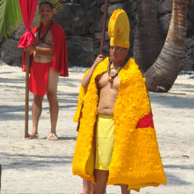 Man in feathered cloak and helmet of the aliʻi (royalty) walks in the Royal Grounds