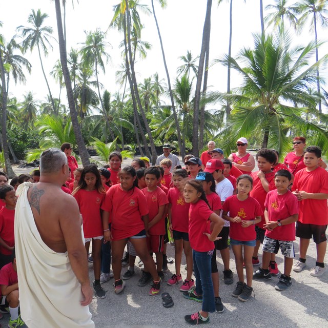 A man in traditional clothing greets a group of school children wearing red t-shirts.