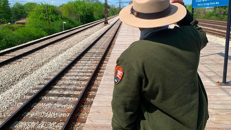 A ranger stands on a train platform looking away from the camera and instead at the tracks.