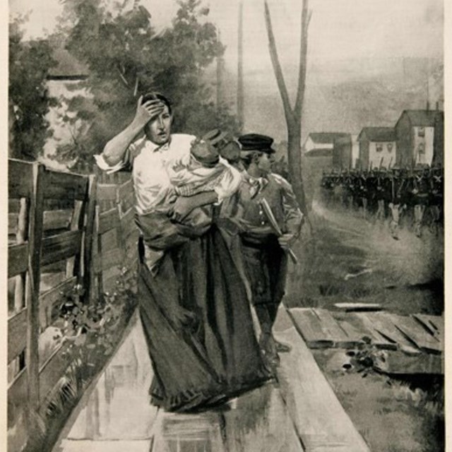 A woman carries a babies in her arms and boy is close behind looking at soldiers in street.