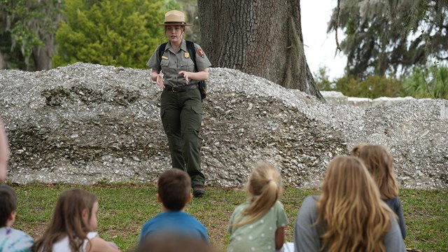 A ranger stands in front of a fort talking to a group of children.