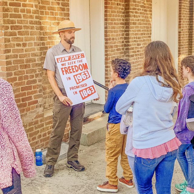 A park ranger holding a sign in front of a brick building, talking to a group of children. 