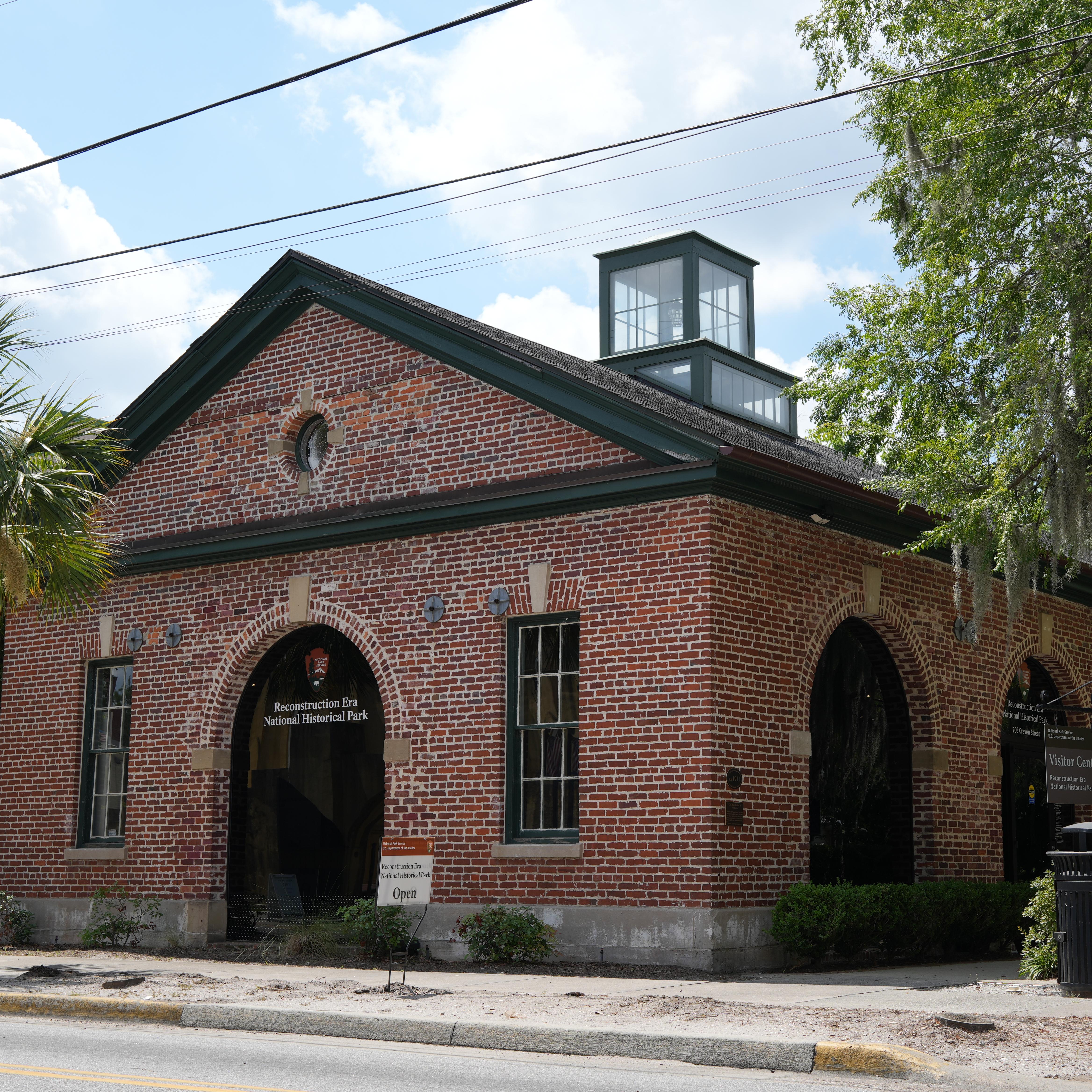 A single story brick building with large arched windows under oak trees and a blue sky.
